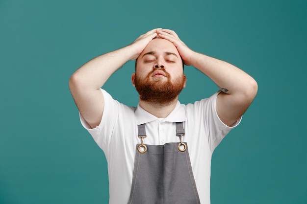 Tired young male barber wearing white shirt and barber apron keeping hands on head with closed eyes isolated on blue background