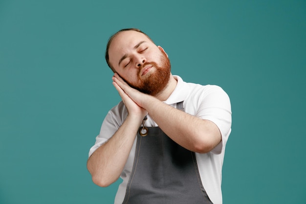 Tired young male barber wearing white shirt and barber apron showing sleep gesture with closed eyes isolated on blue background