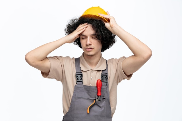 Stanco giovane operaio edile maschio che indossa uniforme e casco di sicurezza che tocca la testa togliendosi il casco con gli occhi chiusi con cacciavite e pinze in tasca isolati su sfondo bianco