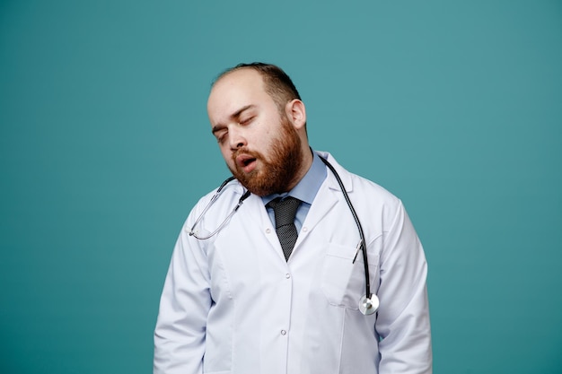 Tired young male doctor wearing medical coat and stethoscope around his neck bending head to side with closed eyes isolated on blue background