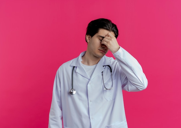 Усталый молодой врач-мужчина в медицинском халате и стетоскопе, держащий нос с закрытыми глазами, изолирован на розовой стене с копией пространства