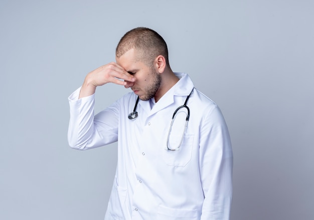 Усталый молодой мужчина-врач в медицинском халате и стетоскопе на шее, держащий нос с закрытыми глазами на белом фоне с копией пространства
