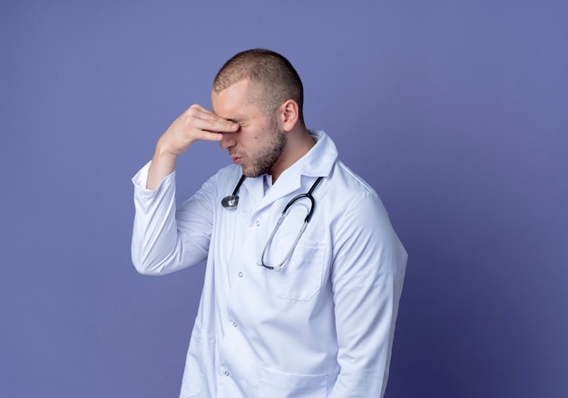 Tired young male doctor wearing medical robe and stethoscope around his neck putting hands on eyes isolated on purple background with copy space