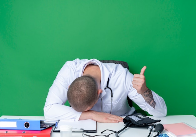 Tired young male doctor wearing medical robe and stethoscope sitting at desk with work tools putting hand on desk and head on hand and showing thumb up isolated on green background