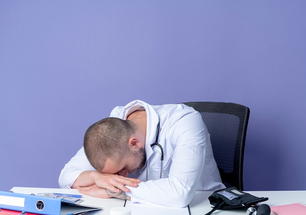 Stanco giovane maschio medico indossando abito medico e stetoscopio seduto alla scrivania con strumenti di lavoro mettendo le mani sulla scrivania e la testa sulle mani isolate su sfondo viola