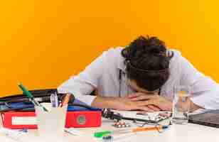 Free photo tired young male doctor with medical glasses wearing medical robe with stethoscope sitting at desk work on laptop with medical tools lowered his head on isolated yellow wall with copy space