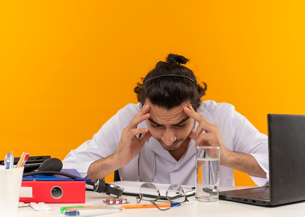 Tired young male doctor with medical glasses wearing medical robe with stethoscope sitting at desk