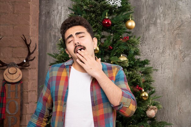 Foto gratuita giovane stanco che sbadiglia davanti all'albero di natale.