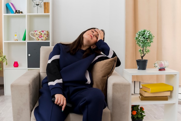 Free photo tired young pretty caucasian woman sitting on armchair in designed living room holding remote control putting hand on head with closed eyes