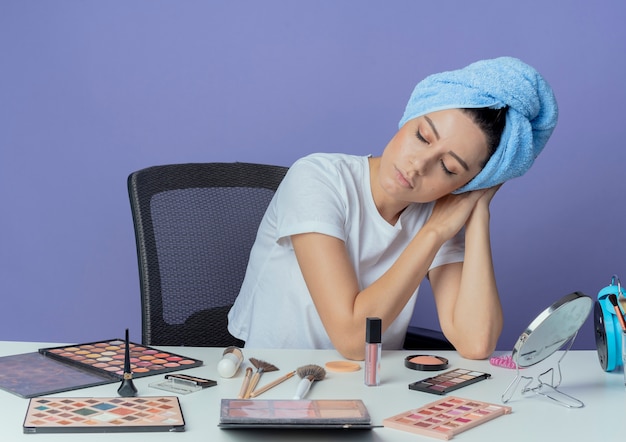 化粧ツールと頭にバスタオルで化粧台に座って、紫色の背景に分離された目を閉じて睡眠ジェスチャーをしている疲れた若いかわいい女の子