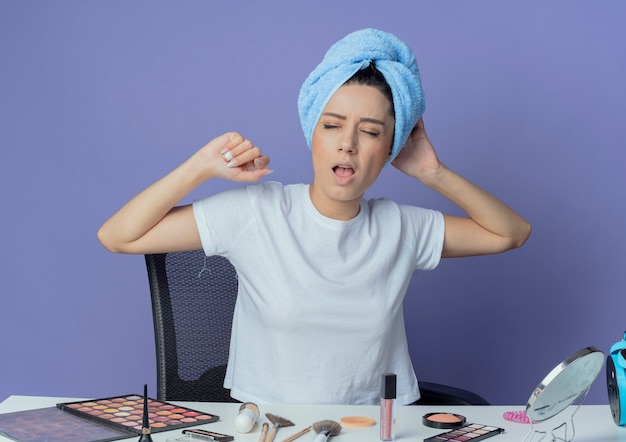 Stanco giovane bella ragazza seduta al tavolo da trucco con strumenti per il trucco e con asciugamano da bagno sulla testa che sbadiglia con gli occhi chiusi e tocca la testa e tiene la mano in aria