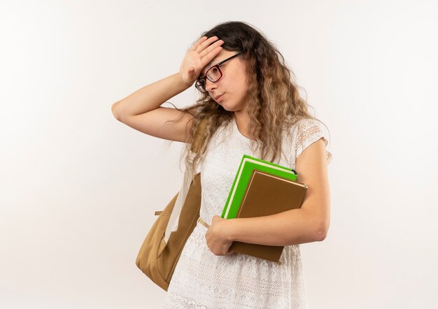 Tired young pretty schoolgirl wearing glasses and back bag holding books looking down putting hand on head isolated on white background with copy space