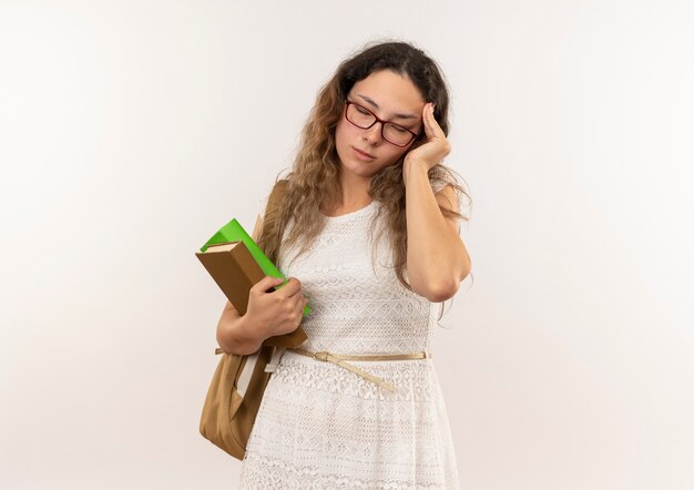 Tired young pretty schoolgirl wearing glasses and back bag holding books putting hand on head with closed eyes isolated on background with copy space