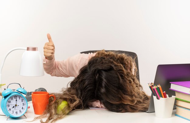 Tired young pretty schoolgirl wearing glasses sitting at desk with school tools putting head on desk and showing thumb up isolated on white background