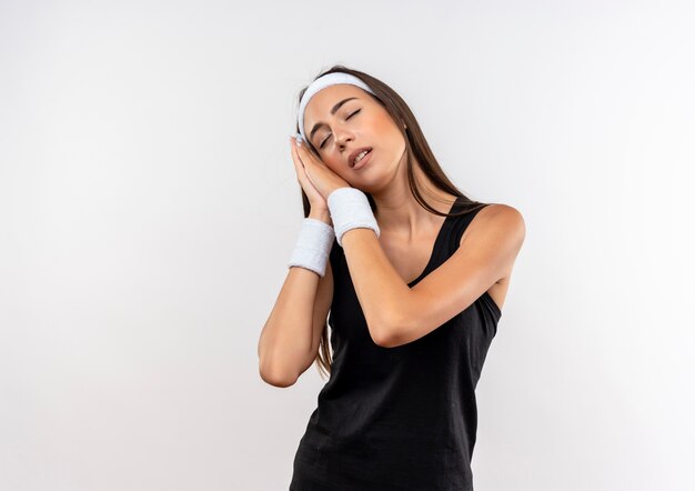 Tired young pretty sporty girl wearing headband and wristband gesturing sleep on white space 