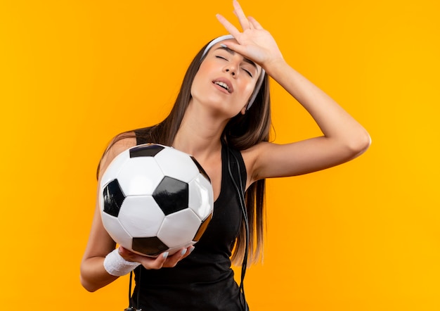Tired young pretty sporty girl wearing headband and wristband holding soccer ball putting hand on head with closed eyes with jumping rope around her neck isolated on orange space