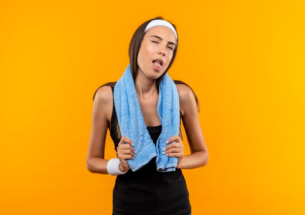 Tired young pretty sporty girl wearing headband and wristband holding towel around her neck showing tongue with one eye closed on orange space