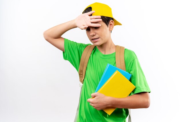 Tired young school boy wearing backpack with cap holding books putting hand on forehead isolated on white wall