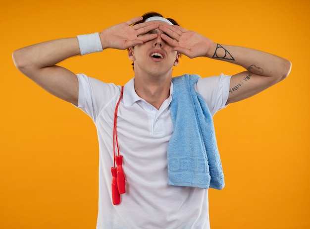 Tired young sporty guy wearing headband with wristband with jump rope and towel on shoulder putting hand on eyes