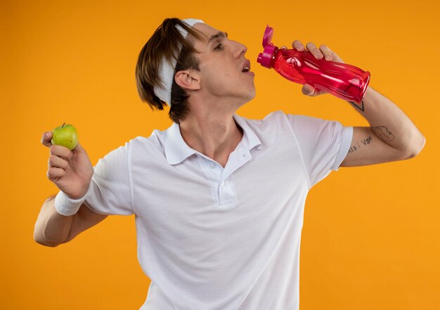 Tired young sporty guy wearing headband and wristband drinks water holding apple isolated on orange wall