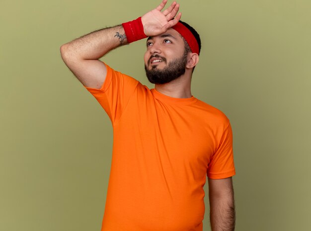 Tired young sporty man looking up wearing headband and wristband wiping forehead with hand isolated on olive green background