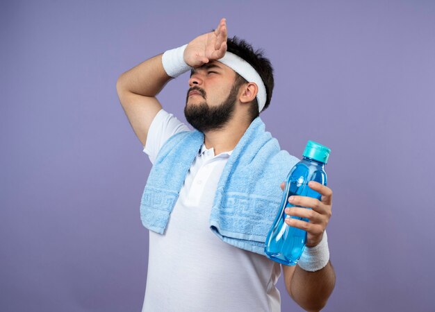 Tired young sporty man wearing headband and wristband holding water bottle with towel on shoulder wiping forehead with hand