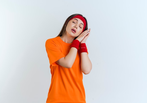 Tired young sporty woman wearing headband and wristbands doing sleep gesture with closed eyes isolated
