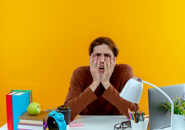 Free photo tired young student boy sitting at desk with school tools covered face with hands on yellow