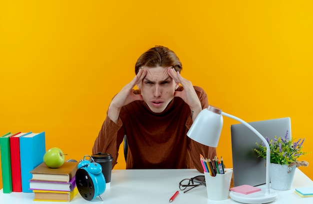 Free photo tired young student boy sitting at desk with school tools putting hands around eyes isolated on yellow wall