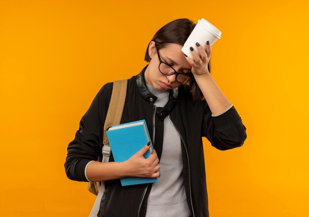 Tired young student girl wearing glasses and back bag holding book touching head with coffee cup with closed eyes isolated on orange background