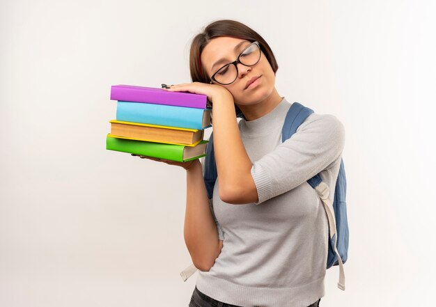 眼鏡と本を保持し、白い背景で隔離の睡眠を試みているそれらに頭を置くバックバッグを身に着けている疲れた若い学生の女の子