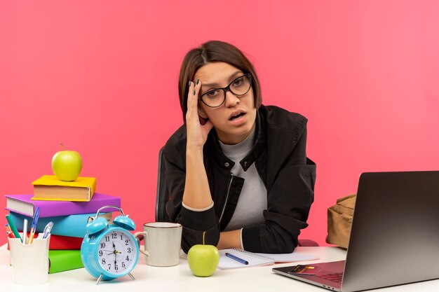 Tired young student girl wearing glasses sitting at desk with university tools putting hand on forehead isolated on pink background