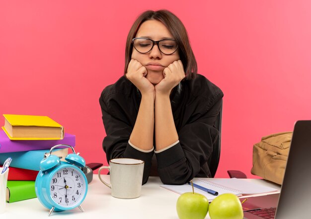 Tired young student girl wearing glasses sitting at desk with university tools putting hands under chin with closed eyes doing homework isolated on pink background