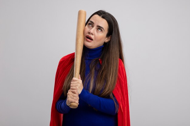 Tired young superhero girl putting baseball bat on cheek isolated on white