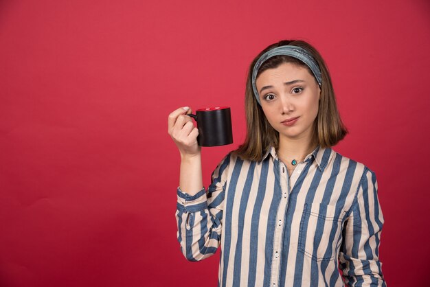 Tired young woman showing cup of coffee and posing to front