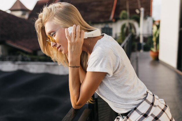 ヘッドフォンで屋外に立っている疲れた若い女性スケートパークで身も凍るように音楽を聴いている壮大なブロンドの女の子