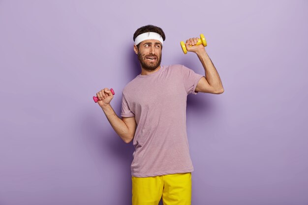 Tiredness and workout concept. Displeased unshaven man raises arms with dumbbells, feels fatigue of long training, dressed in active wear