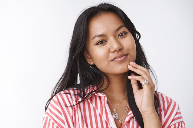 Tnder beautiful and feminine asian female in striped blouse tilting head gentle touching face before putting facial anti acne mask smiling lovely and gazing at camera over white wall