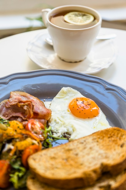 Toast; half fried egg; salad and bacon on gray plate in front of tea cup over the table