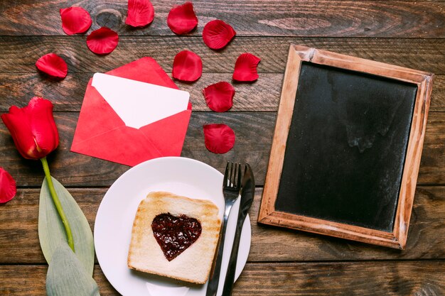 Toast with jam and cutlery on plate near flower, petals, letter and photo frame