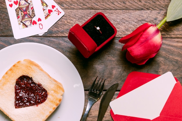 Toast with jam on plate near playing cards, flower, envelope and ring in gift box