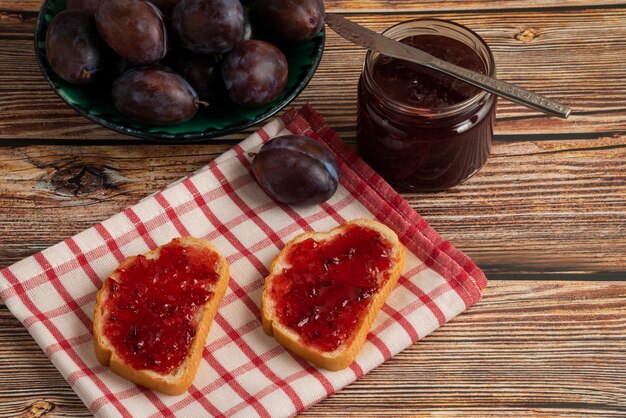 Toastes with plum confiture in a glass jar on a checked towel, top view.