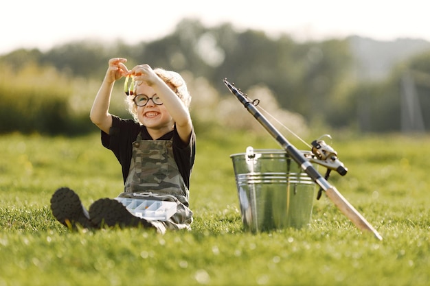 Toddler boy holding a box with a baits for fishing Boy wearing khaki overall Little boy sitting on a grass near the bucket and fishing rod