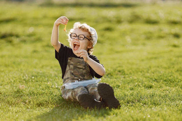 Free photo toddler boy holding a box with a baits for fishing. boy wearing khaki overall. little boy sitting on a grass