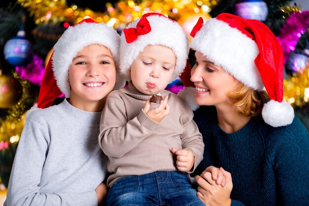 Малыш ест сладкие конфеты в кругу семьи. Улыбается молодая мать с двумя сыновьями в шляпах Санты.