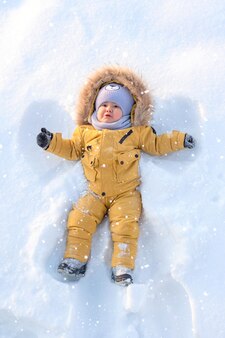 Малыш в желтой зимней одежде лежит на снегу и делает снежного ангела. вертикальное фото