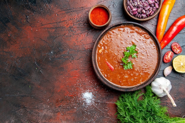 Tomato soup in a brown bowl and different spices garlic lemon and greens on mixed color table