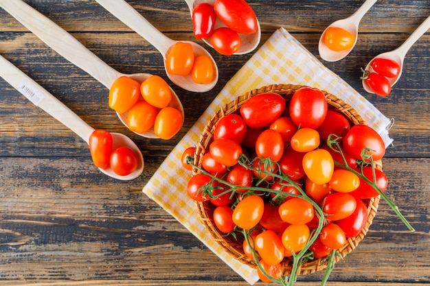 Tomatoes in wooden spoons and basket on wooden and picnic cloth, flat lay.