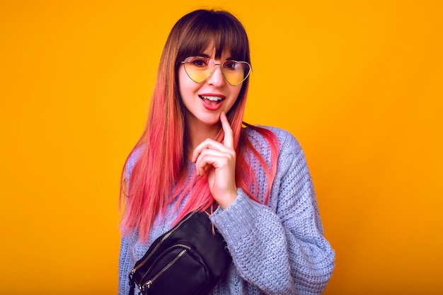 toned portrait of exited happy woman with unusual trendy ombre pink hairs posing at yellow wall, surprised emotions, cozy sweater and vintage sunglasses.
