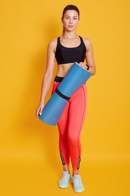 Toned young beautiful dark haired woman athlete wearing sportswear, standing and holding blue fitness mat